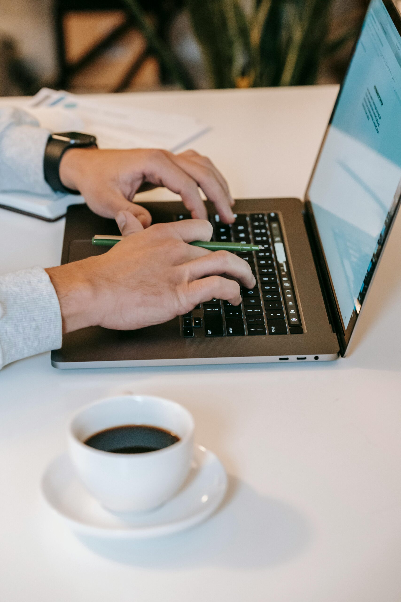 Crop anonymous freelancer sitting at white table with cup of coffee and using laptop in light apartment