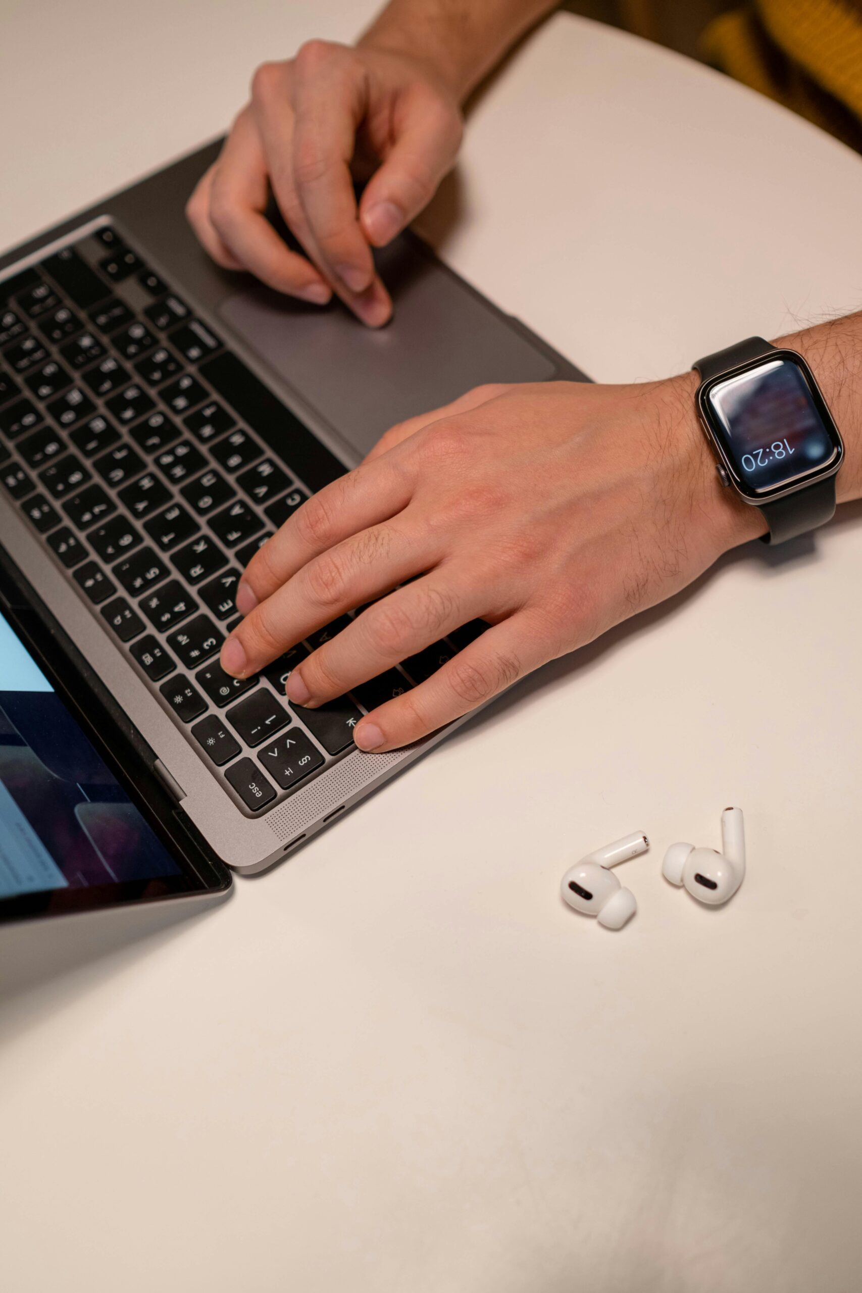 Close-up of a man typing on a laptop while wearing a smartwatch, with earbuds on a table.